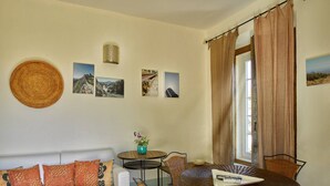 A living room with a white sofa, a small round table, wicker chairs, and a dining table. There are three framed pictures on the wall, a woven wall hanging, and a window with curtains.