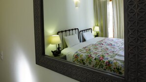 A hotel room with a large mirror, a bed with floral bedding, a nightstand with a lamp, and a window with curtains.