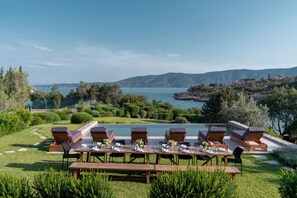 An outdoor dining area with tables and chairs, overlooking a body of water and hills. - Luxury beachfront villa Aria - pool, tennis, sunset views & seaside adventures. (Porto Heli, Argolida)