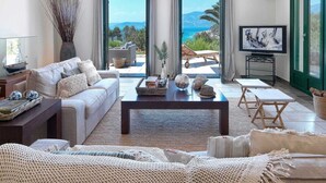 Uma sala de estar ampla com um sofá grande, uma mesa de centro e uma televisão. O quarto tem vista para o mar e para as montanhas através de grandes portas de vidro.