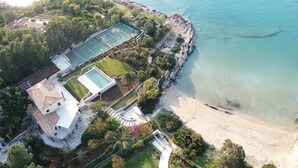 Vista aérea de uma propriedade costeira com quadra de tênis, piscina e acesso à praia.