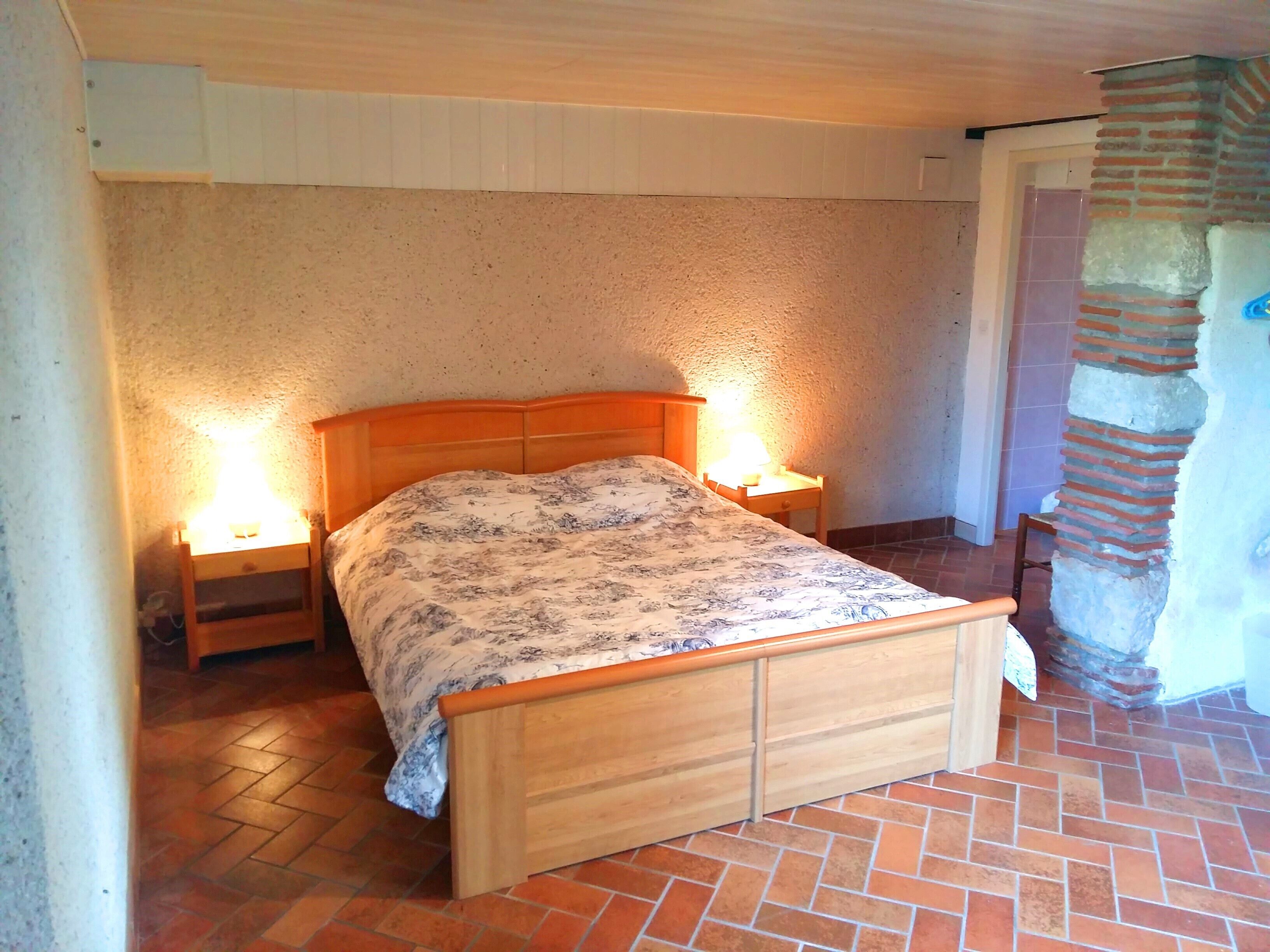 Una cama individual con cabecera y piecera de madera, flanqueada por dos mesitas de noche con lámparas, en una habitación con suelo de baldosas de barro y pared con textura.