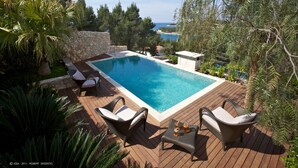 Une piscine rectangulaire avec un mur en pierre, entourée de fauteuils de jardin en osier et d’une terrasse en bois.