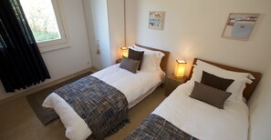 Une chambre d’hôtel avec deux lits, chacun muni d’une table de chevet et d’une lampe. Il y a une fenêtre avec des rideaux, une porte et deux tableaux accrochés au mur.