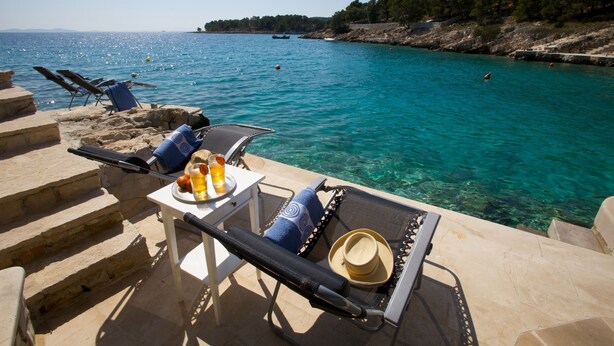 Un espace extérieur ensoleillé, aménagé avec des fauteuils confortables et une table où sont disposés des boissons et un chapeau, offrant une vue imprenable sur une mer bleue et une côte rocheuse.