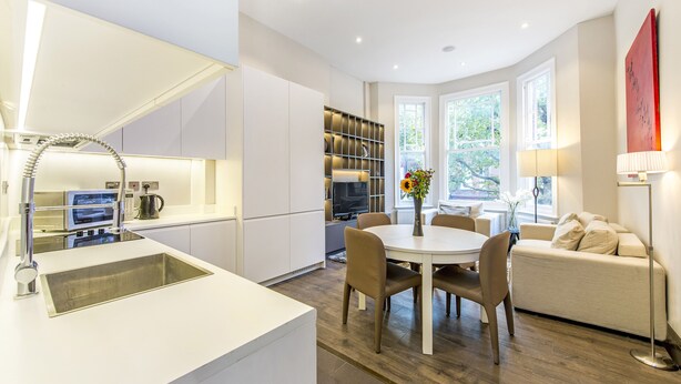 A modern kitchen with a built-in oven, a sink, and a dining area with a round table and chairs. - The One Kensington (London)