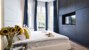 A modern bedroom with a large bed, a TV mounted on the wall, and a window with curtains. - The One Kensington (London)