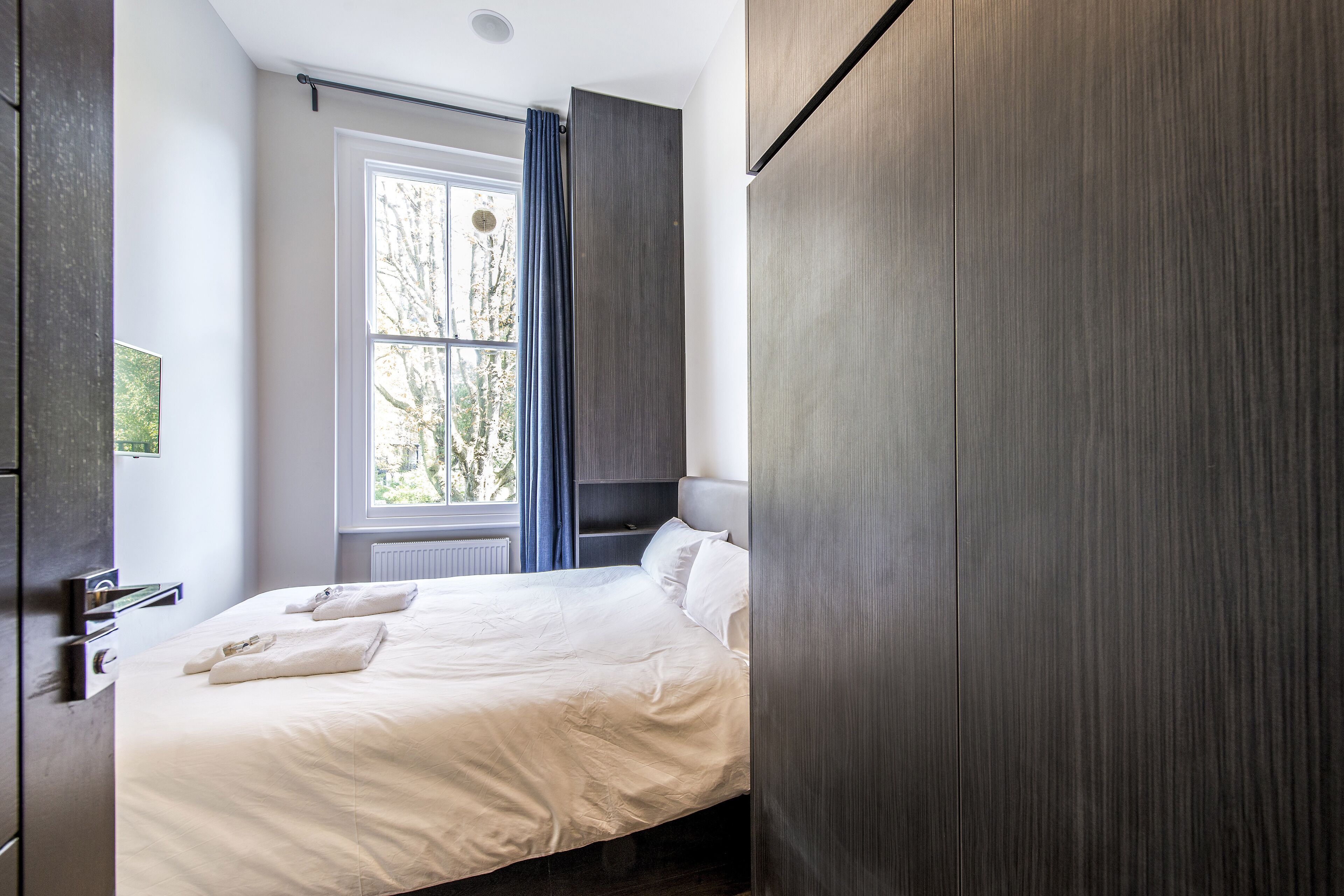 A modern bedroom with a large bed, a window with curtains, and a dark wooden wardrobe.