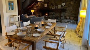Un coin repas avec une table en bois dressée pour six personnes, une cheminée en pierre et un escalier menant à un niveau supérieur.