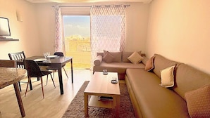 A living room with a sofa, coffee table, dining table, chairs, and a view through a window. - City center, air-conditioned apartment in secure residence with swimming pool. (Marrakech)