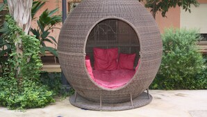 A spherical wicker chair with a red cushion, placed outdoors with plants and a building in the background. - City center, air-conditioned apartment in secure residence with swimming pool. (Marrakech)