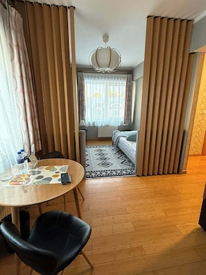 A hotel room with a wooden floor, a round table with chairs, a bed with a patterned rug, and a view through a window with curtains.