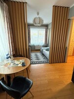 Hotellihuone, jossa on puulattia, pyöreä pöytä tuoleineen, kuviollinen matto sängyn alla ja näkymä ikkunasta, jossa on verhot.
