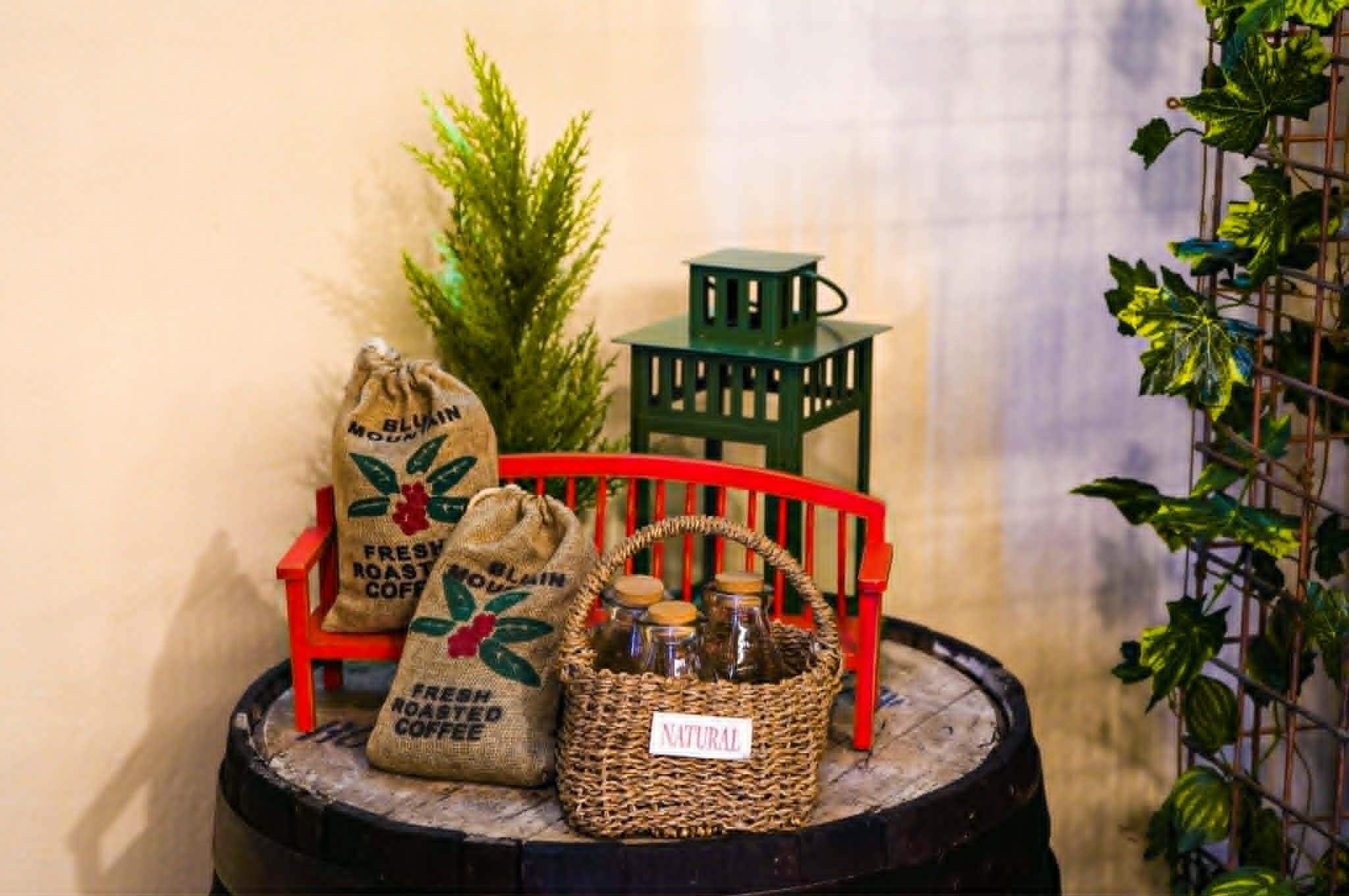 A rustic setting with a burlap sack labeled 'FRESH ROASTED COFFEE,' a wicker basket with jars, a small green lantern, and a red chair.