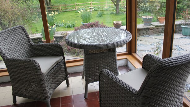A room with wicker furniture and a glass table, overlooking a garden with a wooden fence and potted plants. - Gorgeous Lakeland cottage with delightful private garden. Free EV charging. (Stainton)