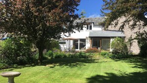 A well-maintained lawn with a stone birdbath, a large tree, and a house with a stone facade and large windows. - Gorgeous Lakeland cottage with delightful private garden. Free EV charging. (Stainton)