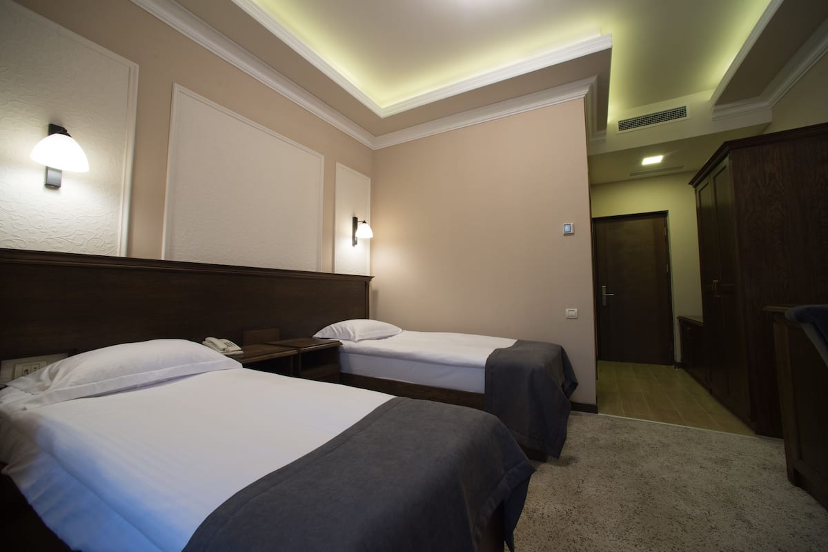Une chambre d’hôtel avec deux lits, une tête de lit en bois, une table de chevet, une lampe fixée au mur et une porte donnant sur un couloir.
