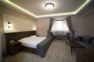 Una habitación de hotel con cama, mesitas de noche, escritorio, silla, sofá y ventana con cortinas.