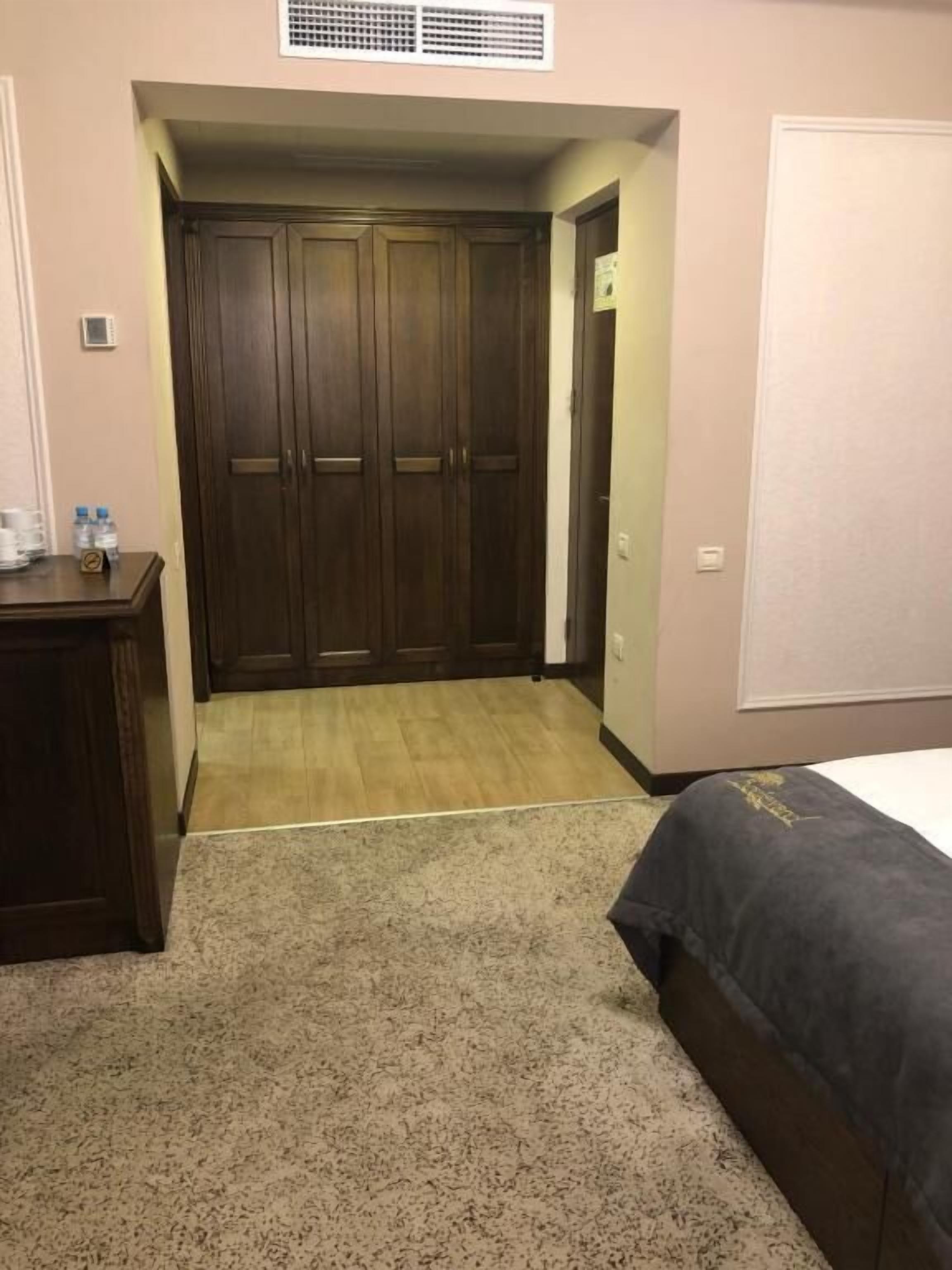 Un dormitorio con un armario de madera, una cama con cubrecama oscuro y suelo alfombrado.