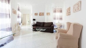 Un salon avec un canapé, un fauteuil et une table basse. On peut voir des rideaux, des œuvres d’art accrochées au mur et une porte au fond.