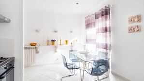 Une cuisine lumineuse aux murs blancs, un coin repas avec des chaises en verre et une table, et une fenêtre agrémentée de rideaux à rayures.