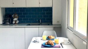 Une cuisine compacte avec des meubles blancs, un pare-feu bleu et un coin repas aménagé sur une petite table près d’une fenêtre.