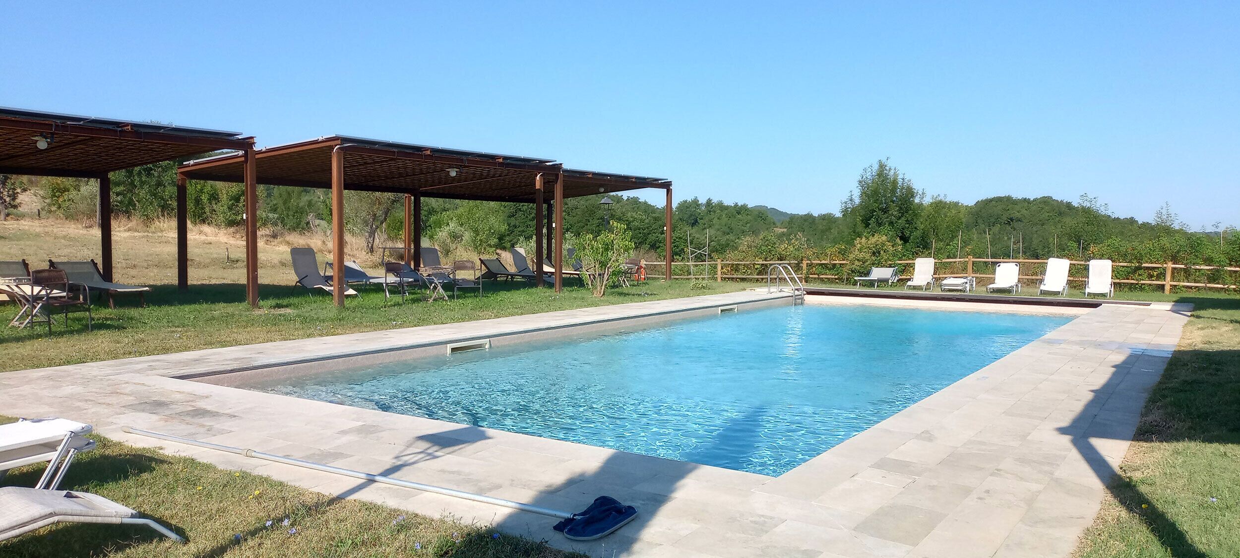Una piscina rectangular con solárium de baldosas, rodeada de sillas de descanso y una pérgola.