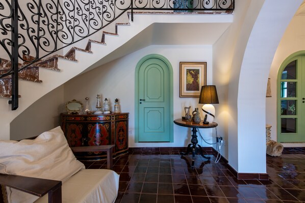 Una escalera con barandales de metal ornamentados, una puerta verde, una mesa con lámpara y un aparador antiguo.