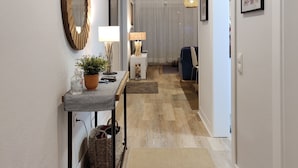 Un pasillo con piso de madera, una consola con lámpara, un espejo y obras de arte enmarcadas en las paredes.