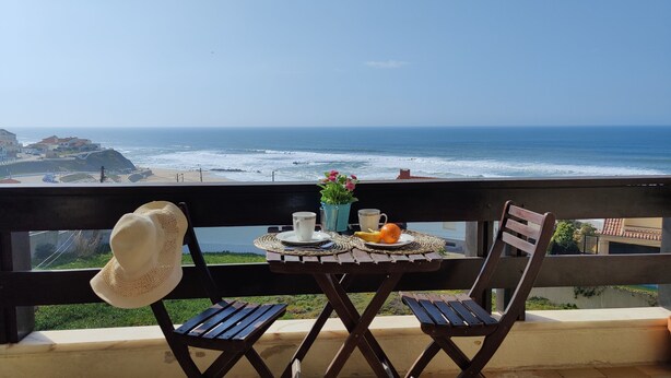Un balcón con una mesa lista para dos personas, con vista a una playa con olas y edificios al fondo.