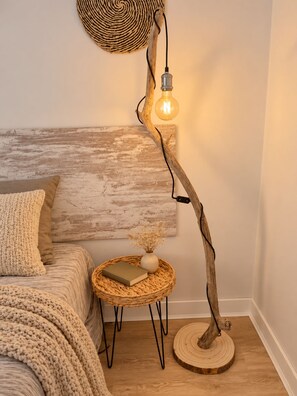 A cozy bedroom with a rustic wooden headboard, a woven side table with a book and vase, and a unique floor lamp with a bulb.