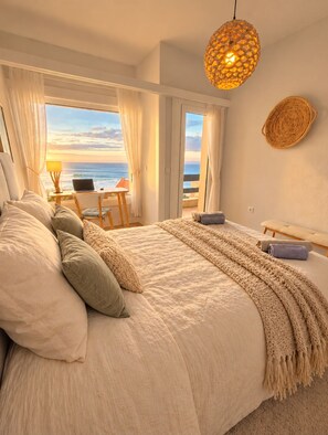 Une chambre avec un grand lit, un ordinateur portable posé sur une table de chevet et une vue sur l’océan.