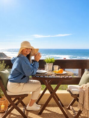 Eine Person sitzt an einem Tisch im Freien mit Blick auf das Meer, einem Blumentopf und einem Korb voller Obst.