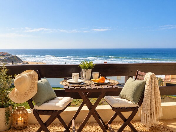A table set on a balcony with a view of the ocean, two chairs, a hat, a candle, and a fruit bowl.