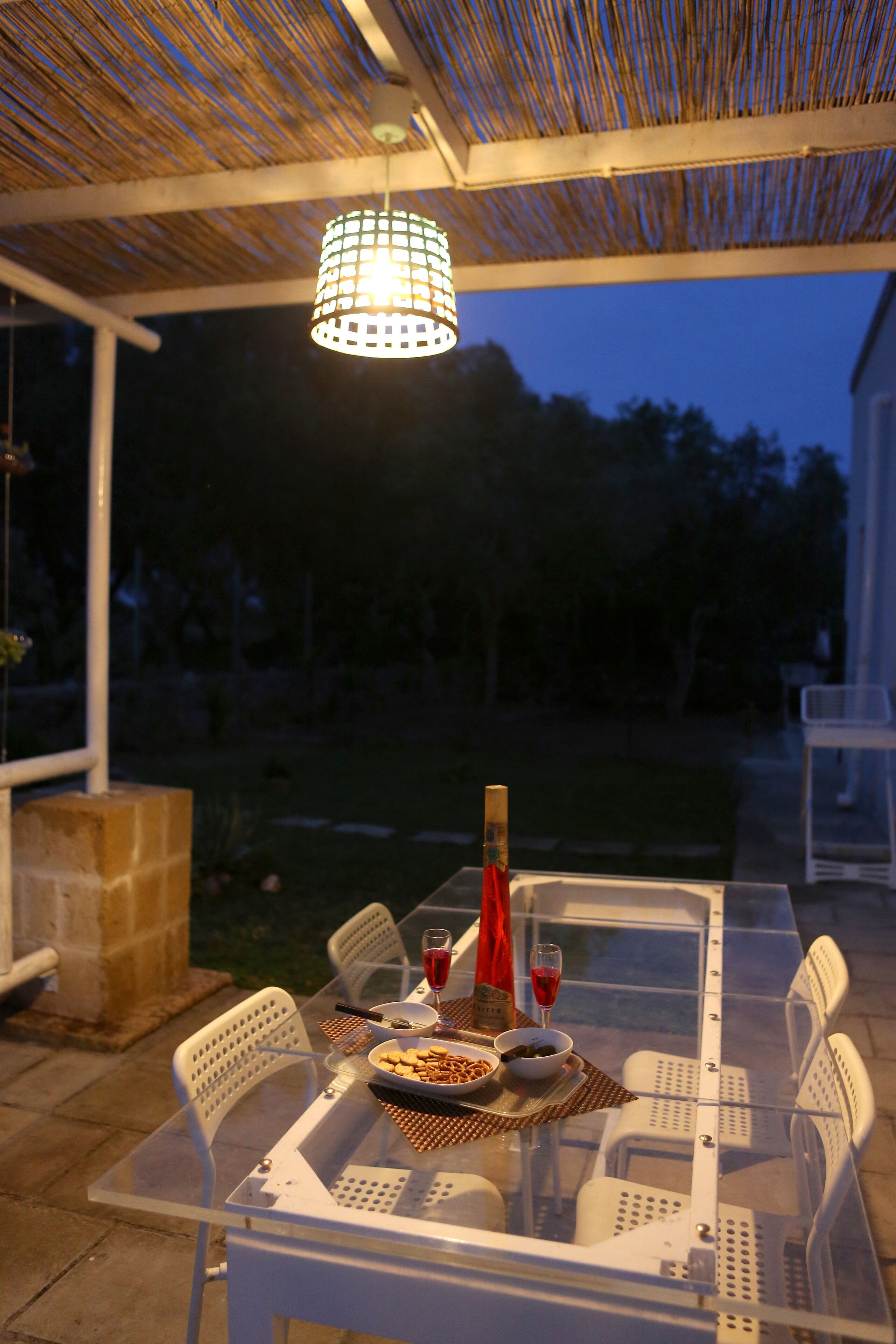 Et udendørs spiseområde med et glasbord og hvide stole, en hængende lampe samt udsigt til baghaven.