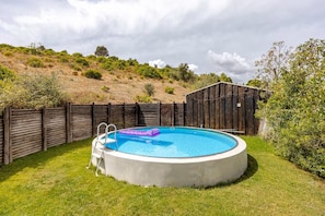 Une piscine circulaire hors sol, dotée d’une échelle et d’un flotteur violet, entourée d’une clôture en bois et d’une pelouse, avec une colline et des arbres en arrière-plan.