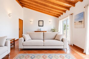 Ein Wohnzimmer mit beiger Couch, Holzbalkendecke, Fliesenboden und Blick durch ein Fenster.