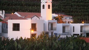 A white building with a clock tower, surrounded by banana plants and a red-roofed structure. - Marina Sol Penthouse <br>- Sea view - Free Wifi (Madalena do Mar)