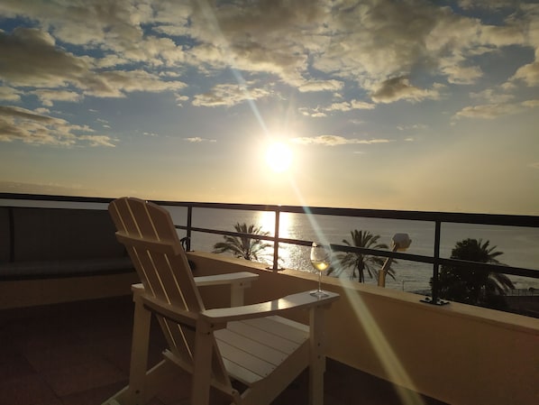 A balcony with a view of the sunset over the sea, a chair, and a glass of wine.