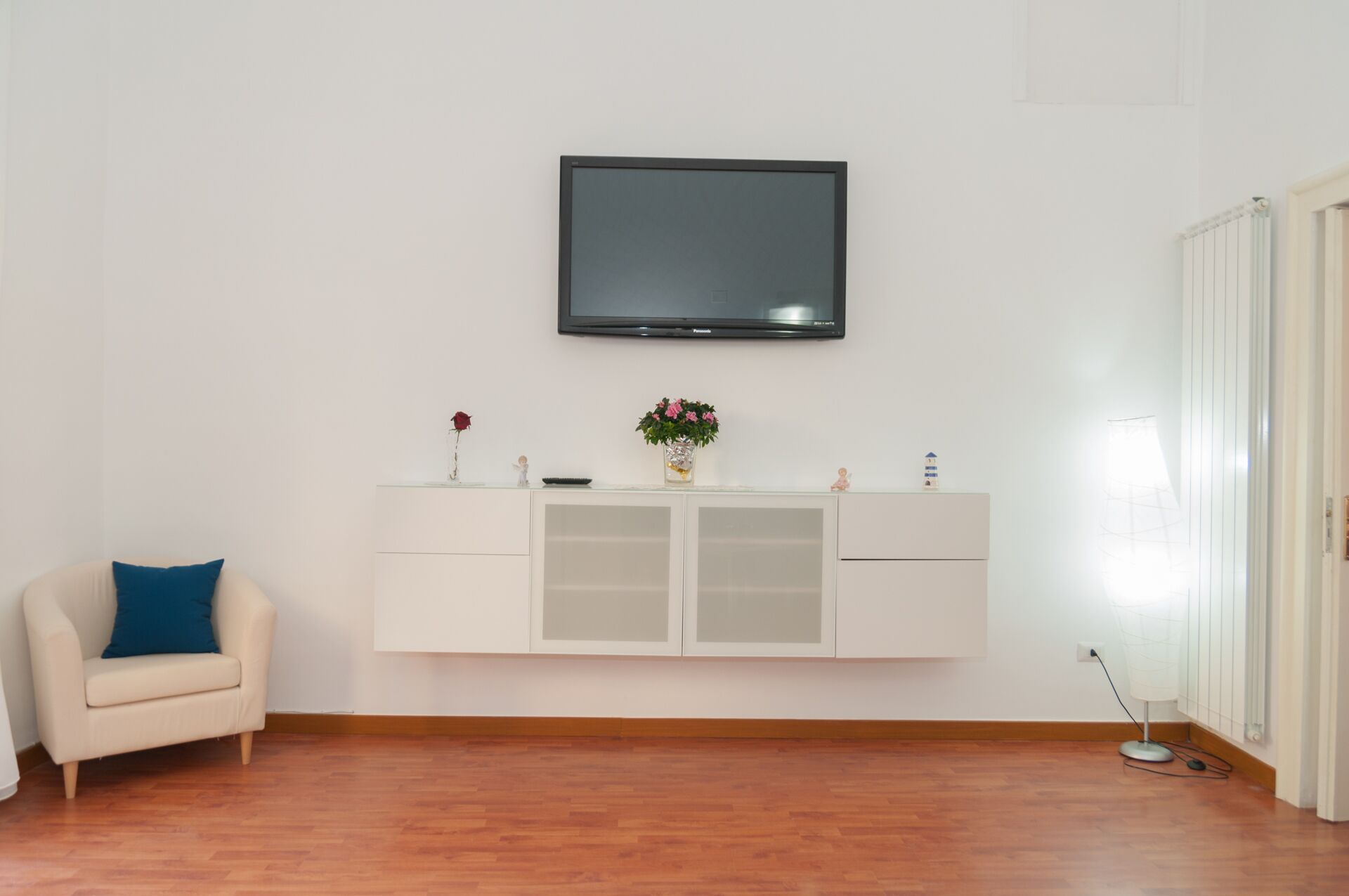 Une pièce avec un téléviseur blanc fixé au mur, un meuble blanc avec des portes en verre, un canapé beige agrémenté d’un coussin bleu et un plancher en bois.