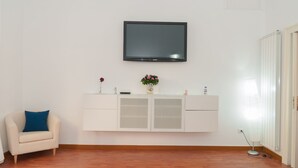 Ein Zimmer mit einer weißen, wandmontierten Fernseher, einer weißen Schranke mit Glastüren, einem beige farbenen Sofa mit blauem Kissen und Holzboden.