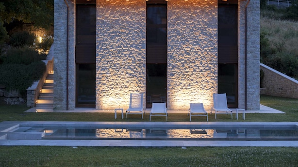 Un edificio moderno con fachada de piedra, grandes ventanales y un área de piscina con sillas de descanso.