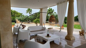A covered outdoor seating area with white plastic furniture, a stone floor, and a view of a garden with palm trees and a fountain. - Es Garrover Es Velar with XXL pool, playground, volleyball, billiards, gym.. (Muro)