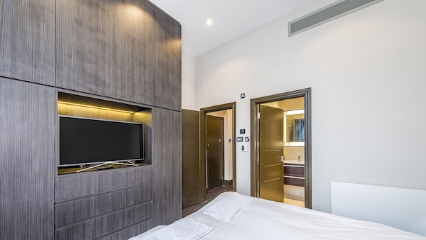 A modern hotel room with a large bed, a flat-screen TV mounted on a wooden panel, a walk-in closet, and a bathroom visible through an open door. - The One Paddington (London)