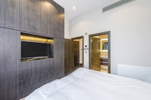 Una habitación de hotel moderna con una cama grande, un televisor de pantalla plana montado en un panel de madera, un vestidor y un baño visible a través de una puerta abierta.