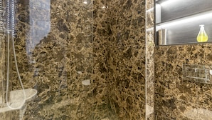 A bathroom with a marble wall, a glass shower enclosure, and a shelf with toiletries. - The One Paddington (London)