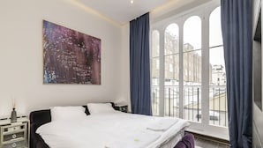 A hotel room with a large bed, a nightstand, a painting, and a view of buildings through a window. - The One Paddington (London)
