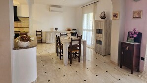 Un comedor amplio con piso de mármol, una mesa de comedor de madera y sillas. Se observa una cocina al fondo con encimera blanca y un equipo de aire acondicionado montado en la pared.