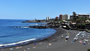 Una playa con gente tomando el sol y nadando, una costa rocosa y edificios al fondo.