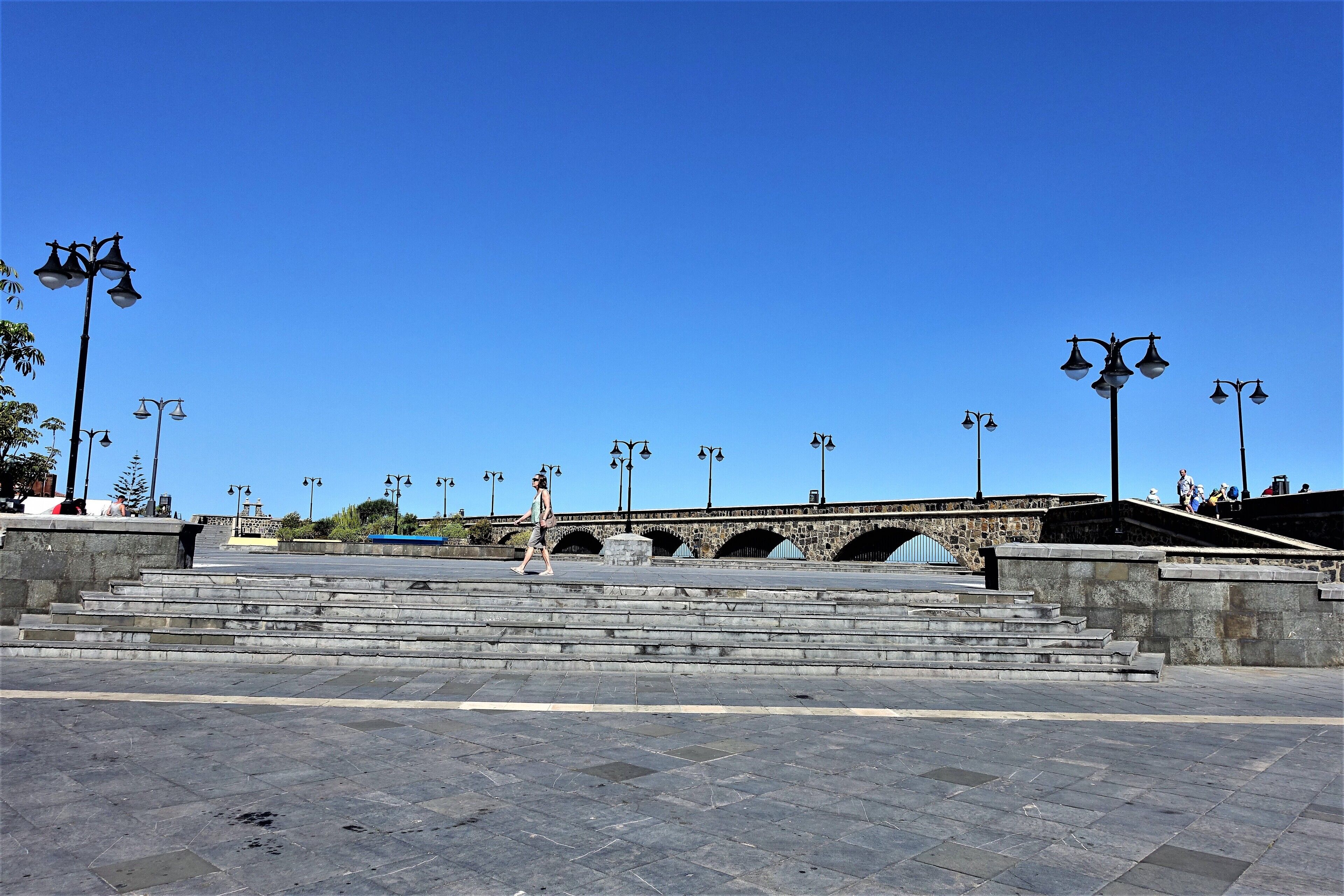 Una plaza pública con escalones de piedra que conducen a un puente, rodeada de farolas espaciadas regularmente.